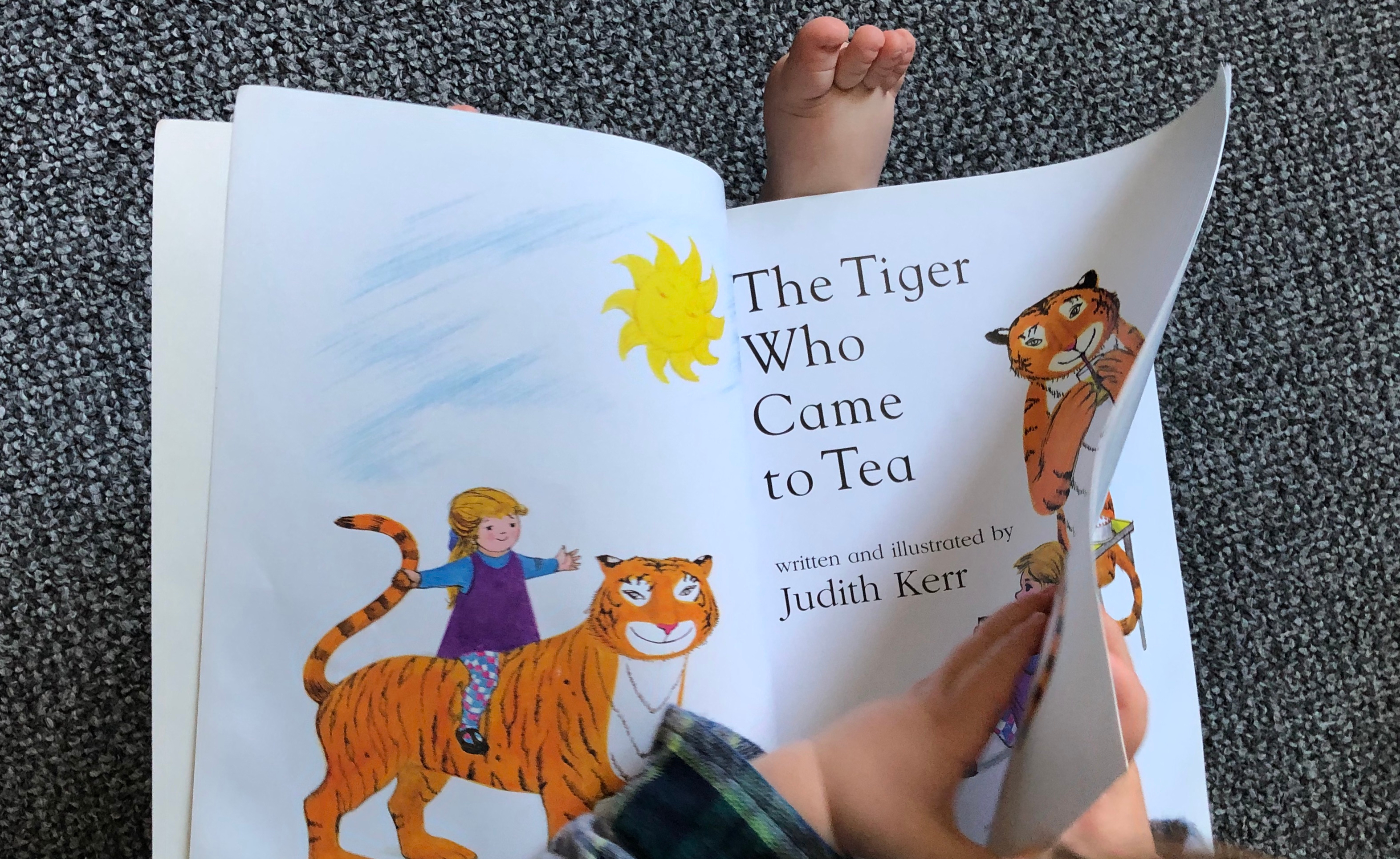 small hands turning the page of a book with a picture of a tiger and the title 'the tiger who came to tea'